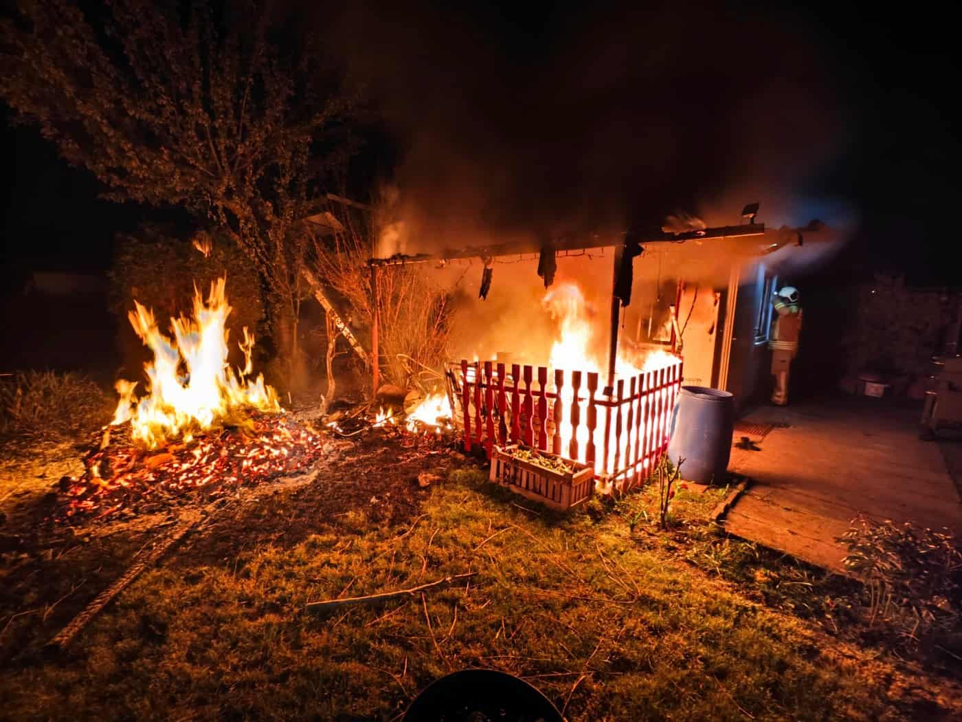 Nächtlicher Einsatz der Feuerwehr Dresden bei einem Gartenlaubenbrand in Nickern. Eine hölzerne Gartenlaube steht in Vollbrand, helle orangefarbene Flammen schlagen aus dem Gebäude und brennen auf der davorliegenden Rasenfläche. Ein Feuerwehrmann in Schutzkleidung ist im Hintergrund bei den Löscharb