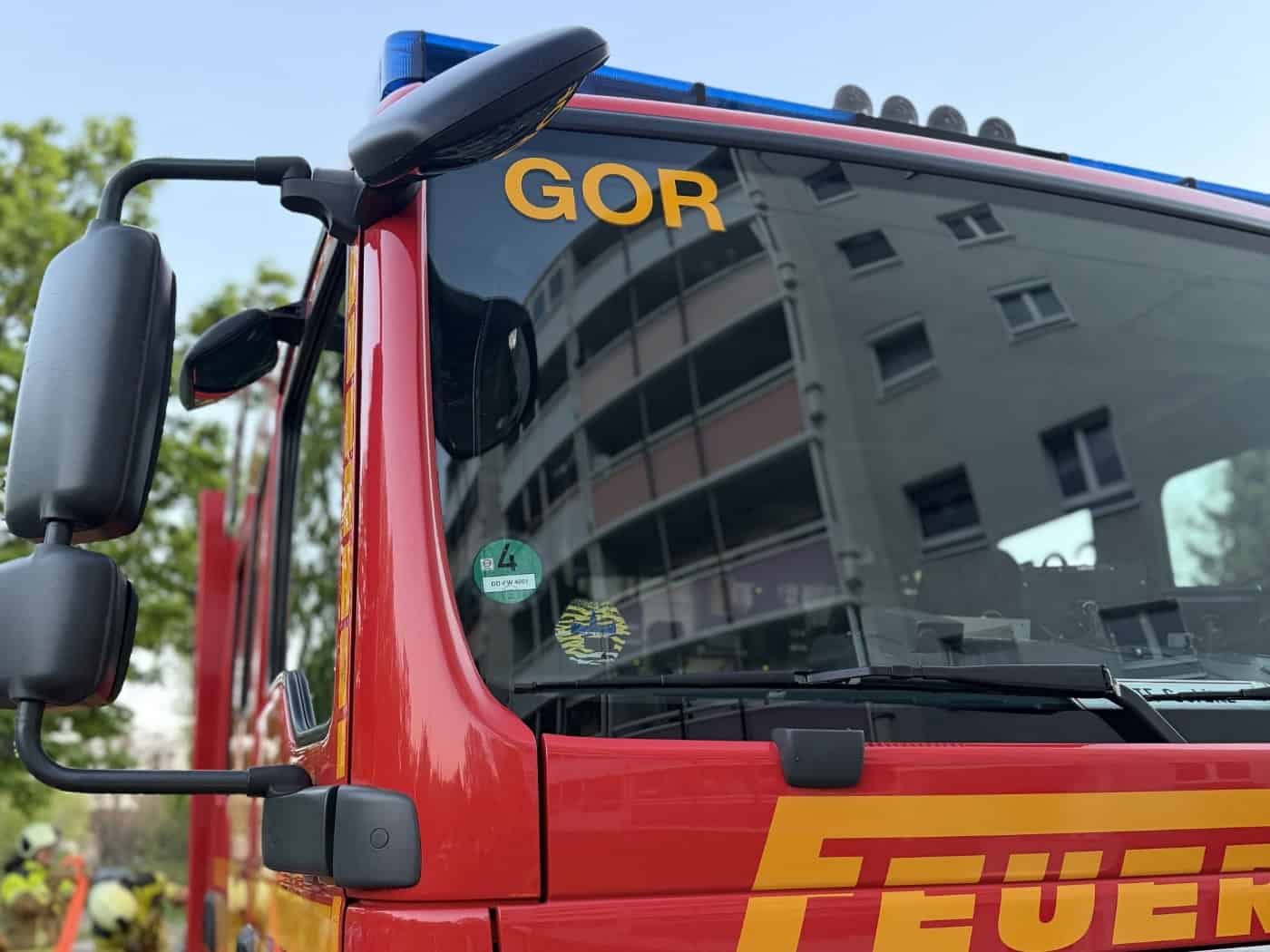 Seitenansicht eines roten Feuerwehr-LKW in Dresden-Gorbitz. In der Windschutzscheibe, die oben mit dem Kürzel „GOR“ markiert ist, spiegelt sich die Fassade eines hellen Plattenbaus. Im Vordergrund ist ein Teil des gelben Schriftzugs „FEUERWEHR“ zu sehen.