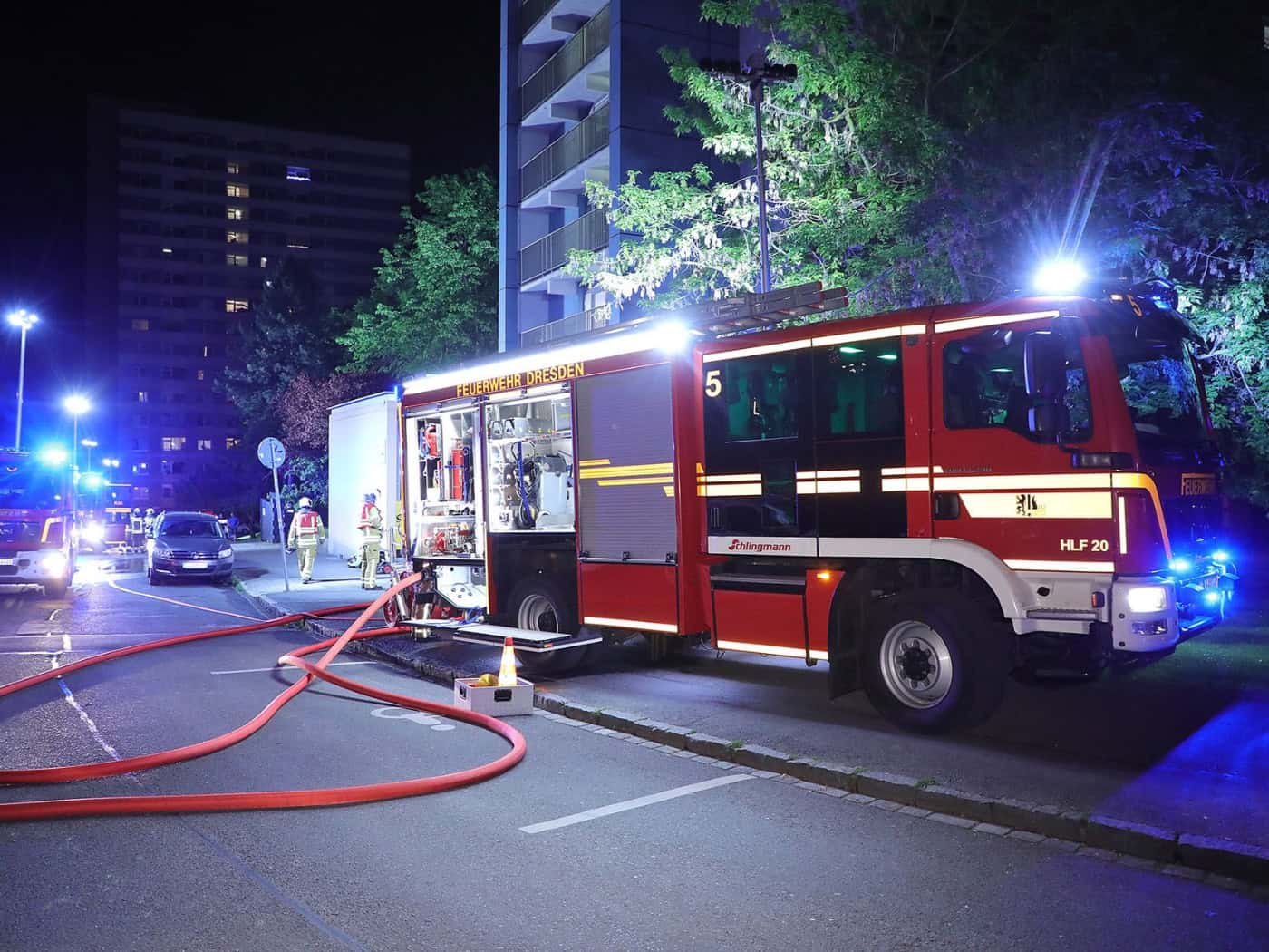 Eine nächtliche Aufnahme eines großen, roten Löschfahrzeugs (HLF 20) der Feuerwehr Dresden, beschriftet mit "FEUERWEHR DRESDEN", im Profil rechts. Es steht auf einer Asphaltstraße, auf der ausgelegte rote Löschschläuche im Vordergrund verlaufen. Im Hintergrund ragt ein hohes Wohnhochhaus auf, dessen
