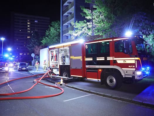 Eine nächtliche Aufnahme eines großen, roten Löschfahrzeugs (HLF 20) der Feuerwehr Dresden, beschriftet mit "FEUERWEHR DRESDEN", im Profil rechts. Es steht auf einer Asphaltstraße, auf der ausgelegte rote Löschschläuche im Vordergrund verlaufen. Im Hintergrund ragt ein hohes Wohnhochhaus auf, dessen