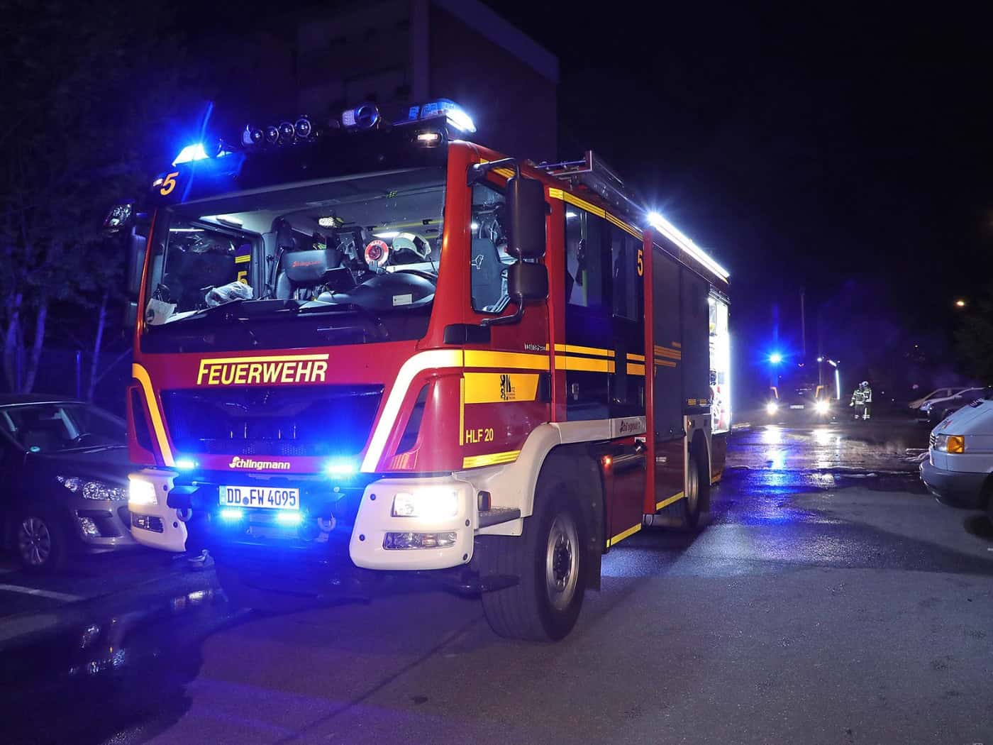 Nächtliche Aufnahme eines modernen, rot-gelben Feuerwehrfahrzeugs (HLF 20) mit eingeschaltetem Blaulicht. Das Fahrzeug mit dem Kennzeichen DD FW 4095 steht auf einer asphaltierten Straße in der Nähe geparkter Autos; im Hintergrund ist ein weiteres Einsatzfahrzeug mit Blaulicht zu sehen.