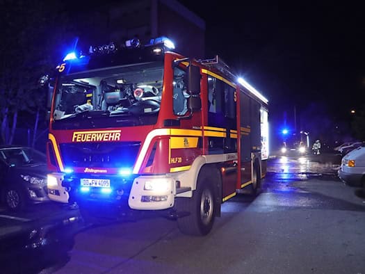Nächtliche Aufnahme eines modernen, rot-gelben Feuerwehrfahrzeugs (HLF 20) mit eingeschaltetem Blaulicht. Das Fahrzeug mit dem Kennzeichen DD FW 4095 steht auf einer asphaltierten Straße in der Nähe geparkter Autos; im Hintergrund ist ein weiteres Einsatzfahrzeug mit Blaulicht zu sehen.