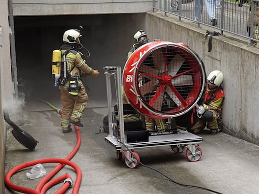Einsatzkräfte der Feuerwehr in voller Montur an einer Tiefgaragenzufahrt. Im Vordergrund steht ein großer, roter Hochleistungsventilator auf einem Rollwagen.