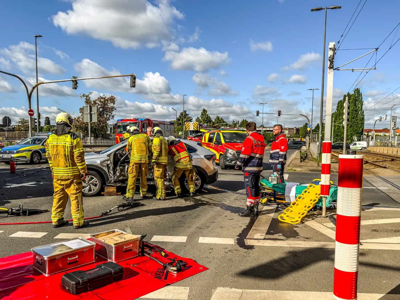 Unfallstelle an einer Straßenkreuzung in Dresden. Mehrere Feuerwehrleute in gelber Schutzkleidung arbeiten mit hydraulischem Rettungsgerät an einem silbernen SUV. Im Vordergrund liegen bereitgestellte Metallkoffer auf einer roten Plane; im Hintergrund sind ein Rettungswagen und ein Notarzteinsatzfah