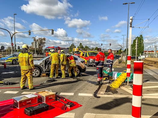 Unfallstelle an einer Straßenkreuzung in Dresden. Mehrere Feuerwehrleute in gelber Schutzkleidung arbeiten mit hydraulischem Rettungsgerät an einem silbernen SUV. Im Vordergrund liegen bereitgestellte Metallkoffer auf einer roten Plane; im Hintergrund sind ein Rettungswagen und ein Notarzteinsatzfah