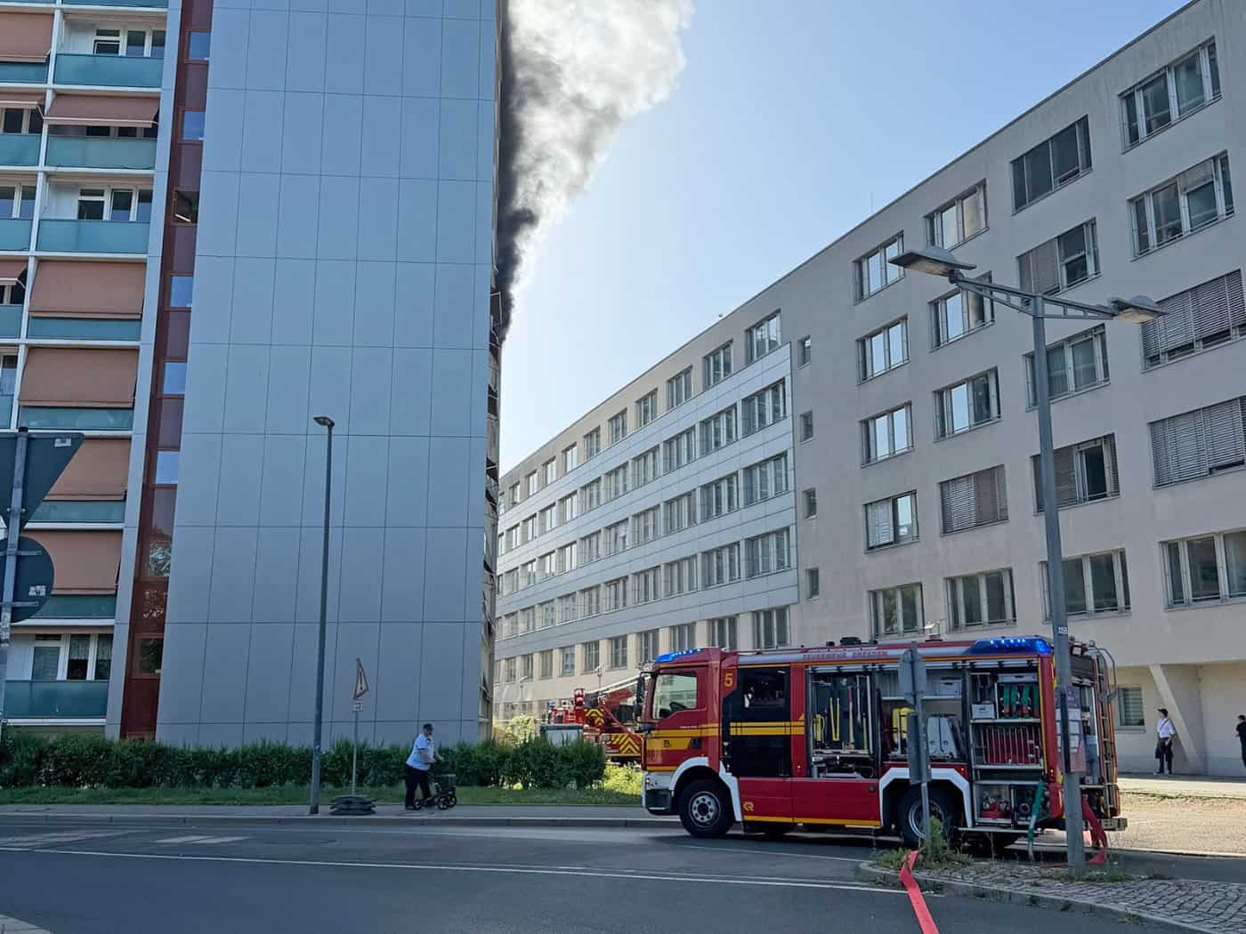 Ein rotes Löschfahrzeug der Feuerwehr Dresden steht auf der Straße vor einem Hochhaus, aus dessen oberen Etagen schwarzer Rauch aufsteigt.