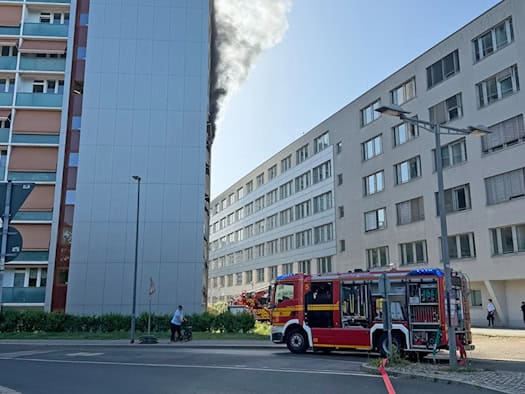 Ein rotes Löschfahrzeug der Feuerwehr Dresden steht auf der Straße vor einem Hochhaus, aus dessen oberen Etagen schwarzer Rauch aufsteigt.