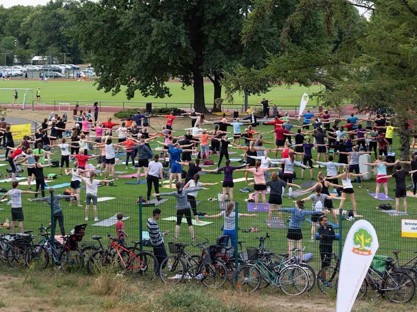 Eine Gruppe von Menschen trainiert gemeinsam auf Gymnastikmatten unter freiem Himmel in einer grünen, sonnigen Parkanlage.