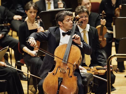 Ein Cellist im dunklen Anzug mit schwarzer Fliege spielt leidenschaftlich auf seinem Instrument inmitten eines Orchesters. Er ist im Fokus der Aufnahme, während im Hintergrund weitere Musiker mit Streichinstrumenten und Notenständer in Unschärfe zu sehen sind.