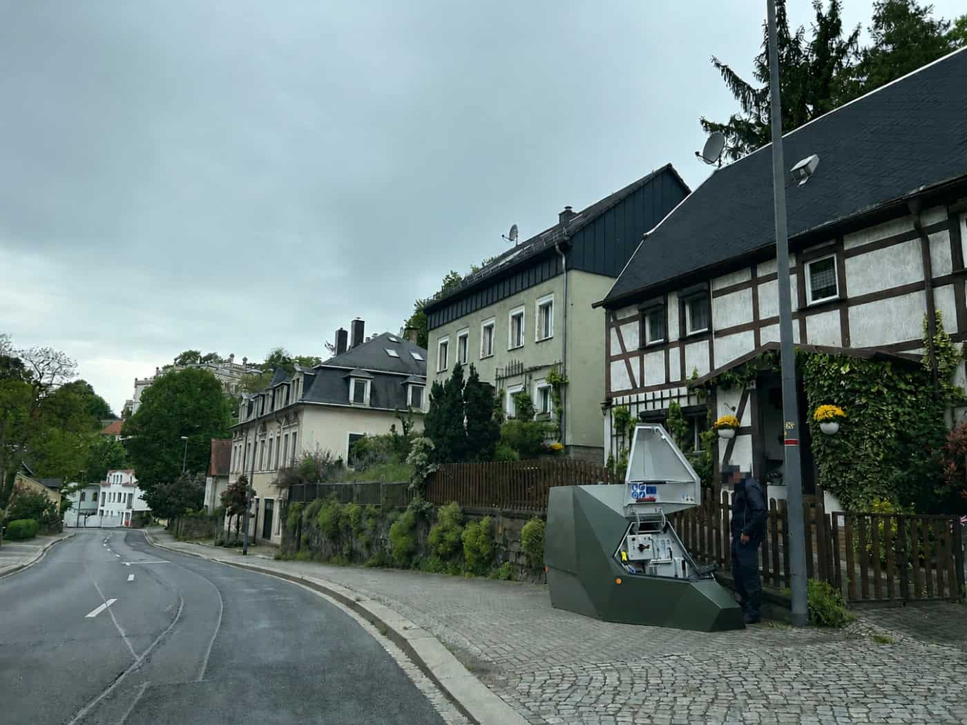 Ein olivgrüner, kantiger Blitzer-Anhänger steht auf einem gepflasterten Gehweg neben einem historischen Fachwerkhaus an einer ansteigenden Straße. Eine Person in dunkler Uniform arbeitet am geöffneten Gehäuse des Geräts.