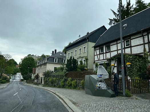 Ein olivgrüner, kantiger Blitzer-Anhänger steht auf einem gepflasterten Gehweg neben einem historischen Fachwerkhaus an einer ansteigenden Straße. Eine Person in dunkler Uniform arbeitet am geöffneten Gehäuse des Geräts.