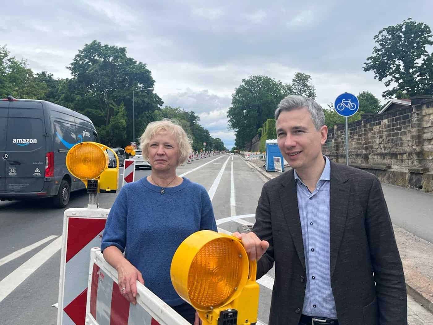 Dresdens Verkehrsbürgermeister Stephan Kühn und Amtsleiterin Simone Prüfer stehen lächelnd hinter einer Baustellenabsperrung mit gelben Warnleuchten an der Stauffenbergallee.