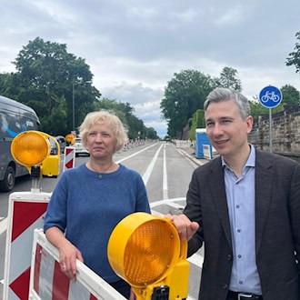 Dresdens Verkehrsbürgermeister Stephan Kühn und Amtsleiterin Simone Prüfer stehen lächelnd hinter einer Baustellenabsperrung mit gelben Warnleuchten an der Stauffenbergallee.