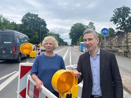 Dresdens Verkehrsbürgermeister Stephan Kühn und Amtsleiterin Simone Prüfer stehen lächelnd hinter einer Baustellenabsperrung mit gelben Warnleuchten an der Stauffenbergallee.