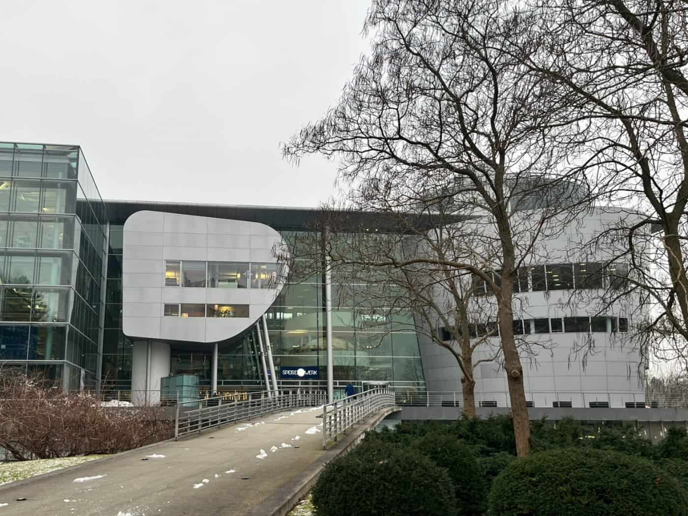 Außenaufnahme der modernen Gläsernen Manufaktur von Volkswagen in Dresden an einem grauen Wintertag. Eine Rampe führt zum Haupteingang, Schnee liegt auf dem Rasen und den Büschen.