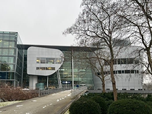 Außenaufnahme der modernen Gläsernen Manufaktur von Volkswagen in Dresden an einem grauen Wintertag. Eine Rampe führt zum Haupteingang, Schnee liegt auf dem Rasen und den Büschen.
