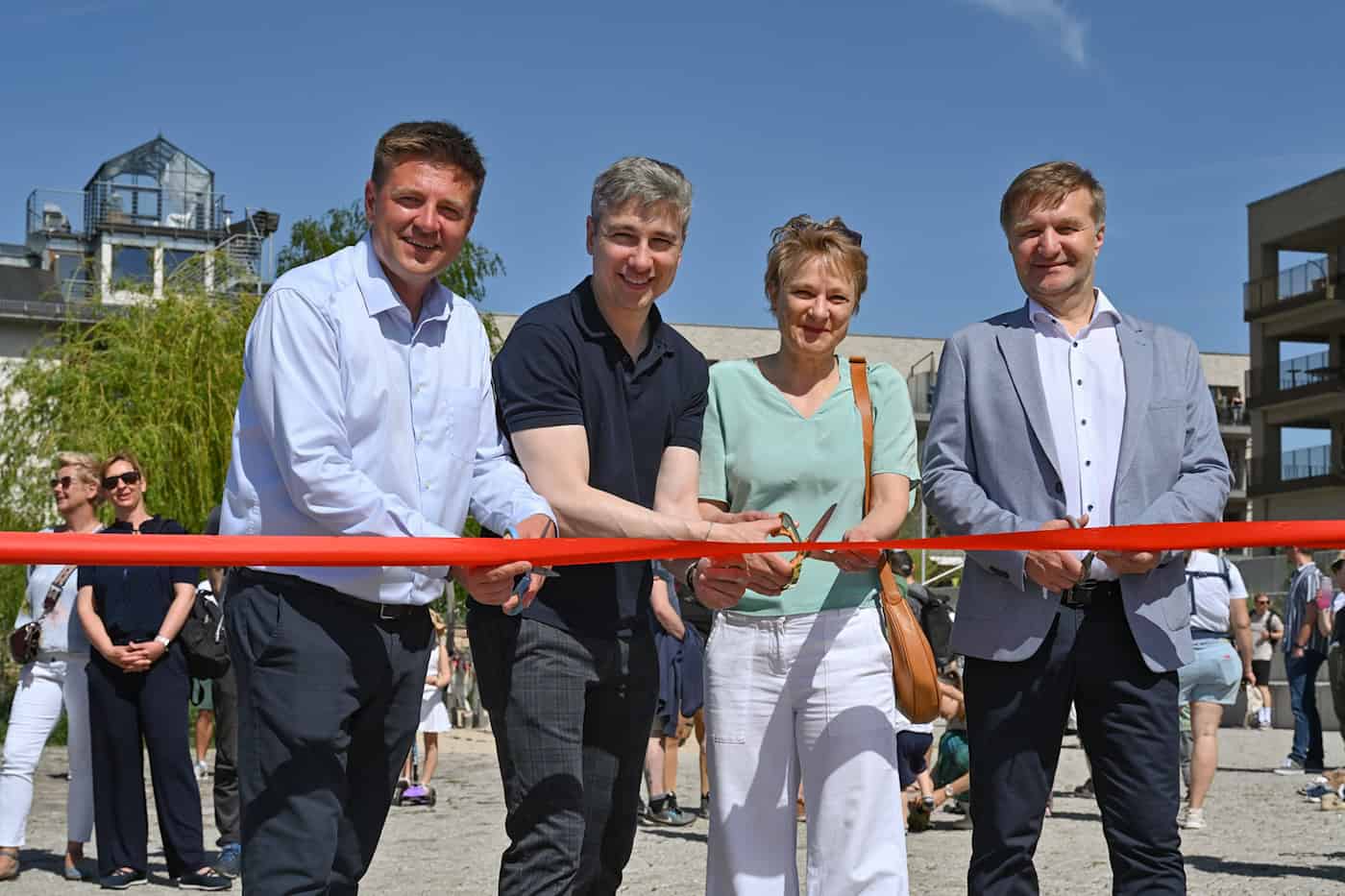 Vier Personen – Architekt Jörn Leschik, Sebastian Forkert von USD Immobilien, Anja Heckmann vom Stadtplanungsamt und Baubürgermeister Stephan Kühn – schneiden gemeinsam ein rotes Band zur Eröffnung der Hafenpromenade in der Dresdner Hafencity durch. Die Szene findet bei strahlendem Sonnenschein vor der Kulisse moderner Wohngebäude und Passanten statt.