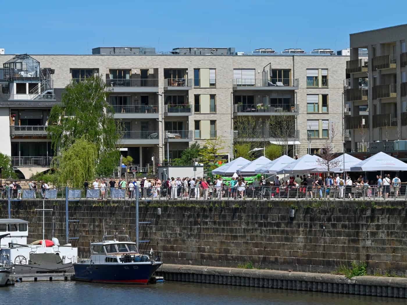 Blick auf die neu eröffnete Hafenpromenade der Hafencity Dresden bei Sonnenschein. Im Vordergrund liegen Boote an einer massiven Steinkaimauer im Hafenbecken. Auf der Promenade flanieren zahlreiche Besucher vorbei an Restaurant-Außenbereichen mit großen weißen Sonnenschirmen. Im Hintergrund erstreck