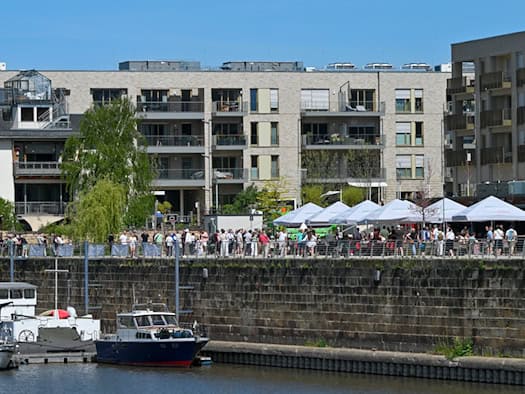 Blick auf die neu eröffnete Hafenpromenade der Hafencity Dresden bei Sonnenschein. Im Vordergrund liegen Boote an einer massiven Steinkaimauer im Hafenbecken. Auf der Promenade flanieren zahlreiche Besucher vorbei an Restaurant-Außenbereichen mit großen weißen Sonnenschirmen. Im Hintergrund erstreck