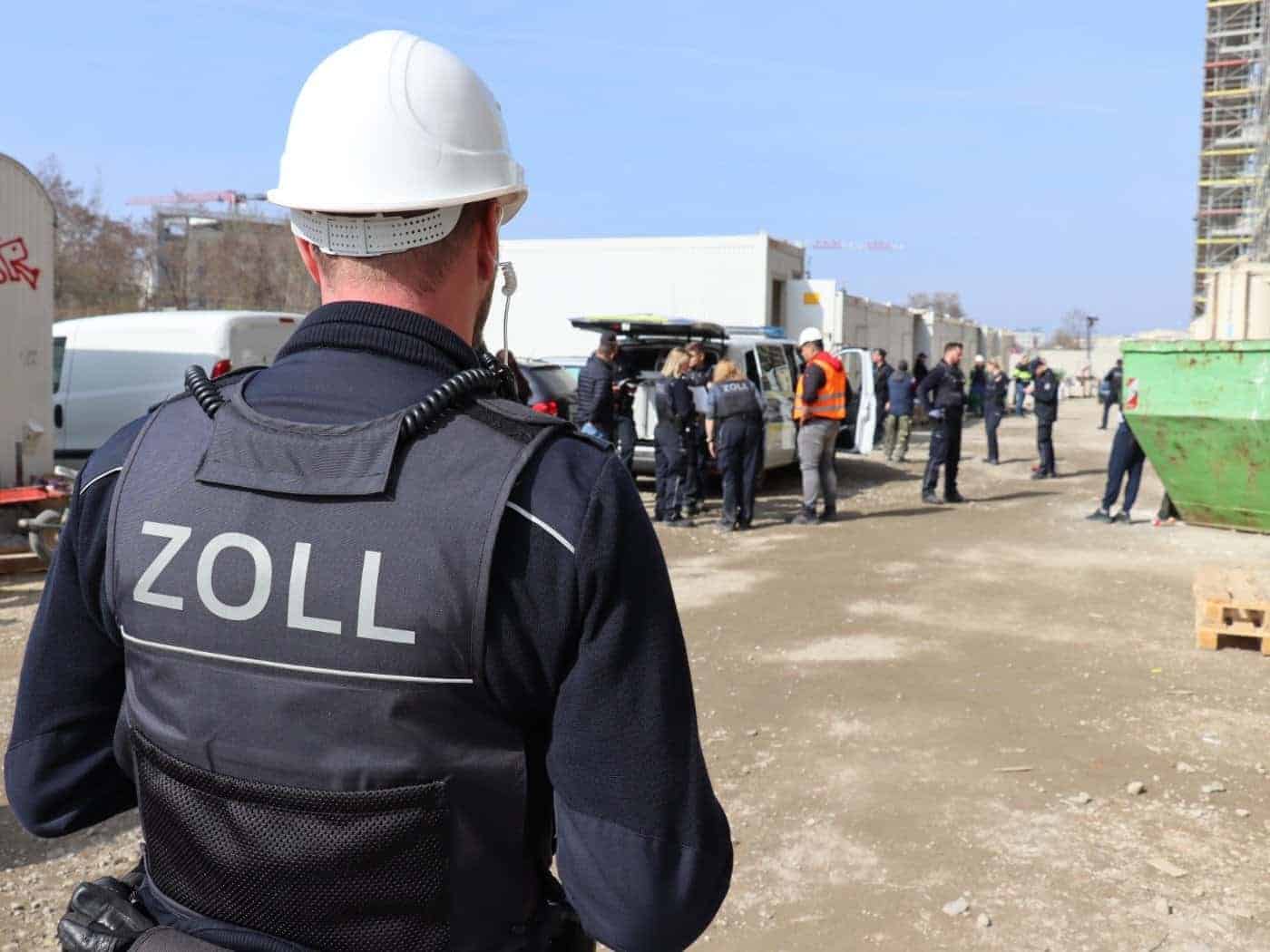 Ein Zollbeamter in dunkelblauer Uniform mit der Aufschrift „ZOLL“ auf dem Rücken steht an einem Tisch und kontrolliert Dokumente. Neben ihm steht ein weiterer Kollege im Profil.
