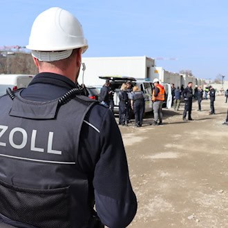Ein Zollbeamter in dunkelblauer Uniform mit der Aufschrift „ZOLL“ auf dem Rücken steht an einem Tisch und kontrolliert Dokumente. Neben ihm steht ein weiterer Kollege im Profil.