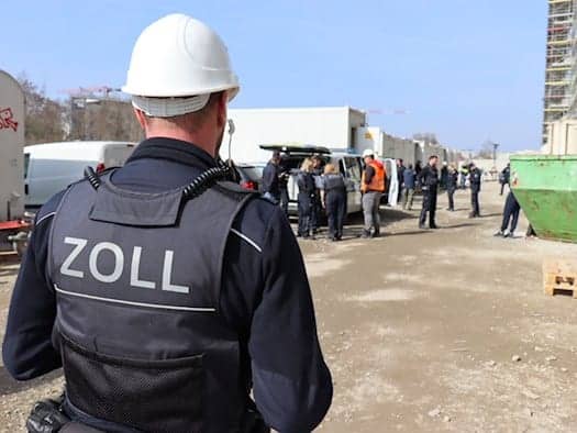 Ein Zollbeamter in dunkelblauer Uniform mit der Aufschrift „ZOLL“ auf dem Rücken steht an einem Tisch und kontrolliert Dokumente. Neben ihm steht ein weiterer Kollege im Profil.