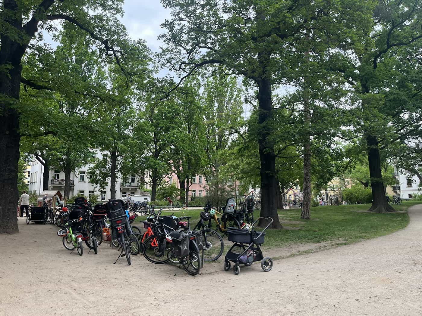 Zahlreiche Fahrräder, Lastenräder und Kinderwagen stehen im Schatten großer, alter Bäume an einer Weggabelung im Hermann-Seidel-Park.