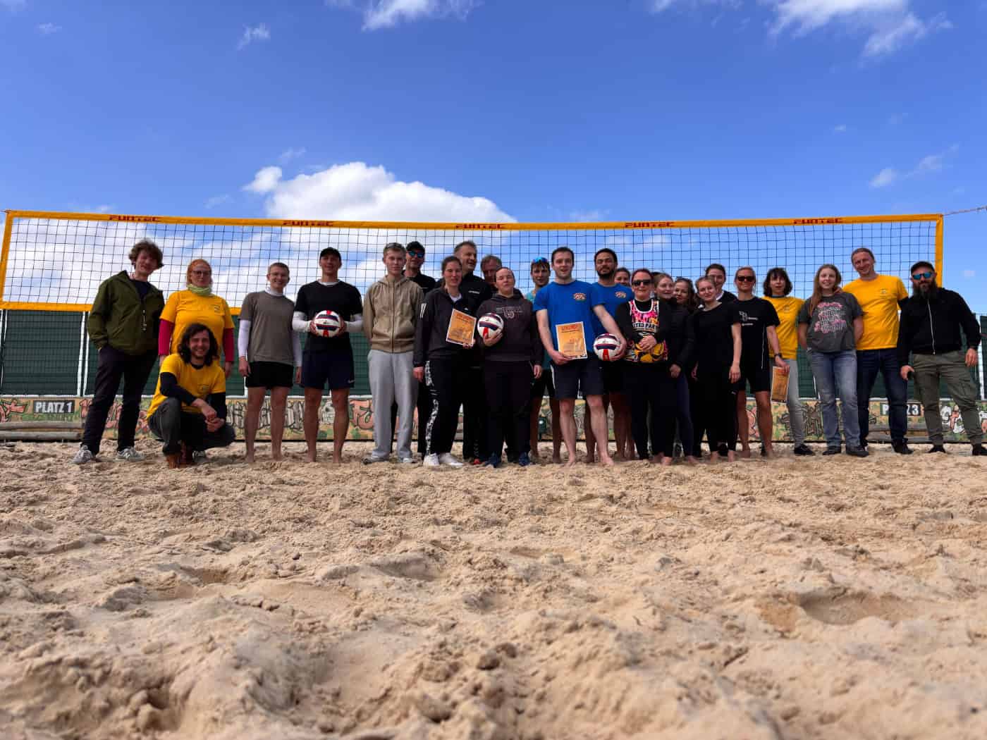 Foto: Zwei Dutzend Spielerinnen und Spieler stehen mit Urkunden und Volleybällen vor einem Beachvolleyballnetz und blauem Himmel.