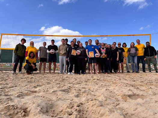 Foto: Zwei Dutzend Spielerinnen und Spieler stehen mit Urkunden und Volleybällen vor einem Beachvolleyballnetz und blauem Himmel.