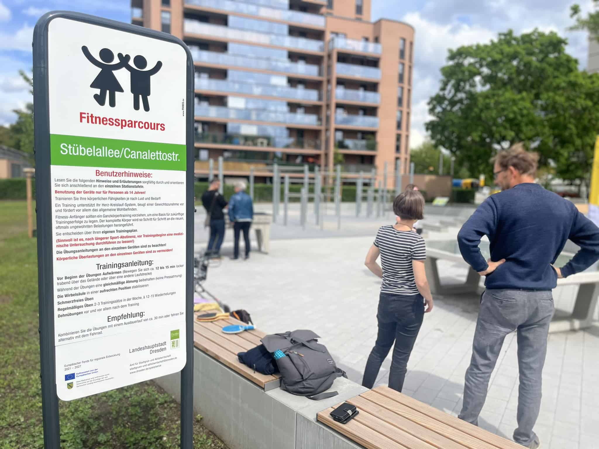 Im Vordergrund steht ein weißes Hinweisschild mit der Aufschrift „Fitnessparcours Stübelallee/Canalettostr.“; im Hintergrund sind Menschen an einer Tischtennisplatte und ein modernes Wohngebäude zu sehen.