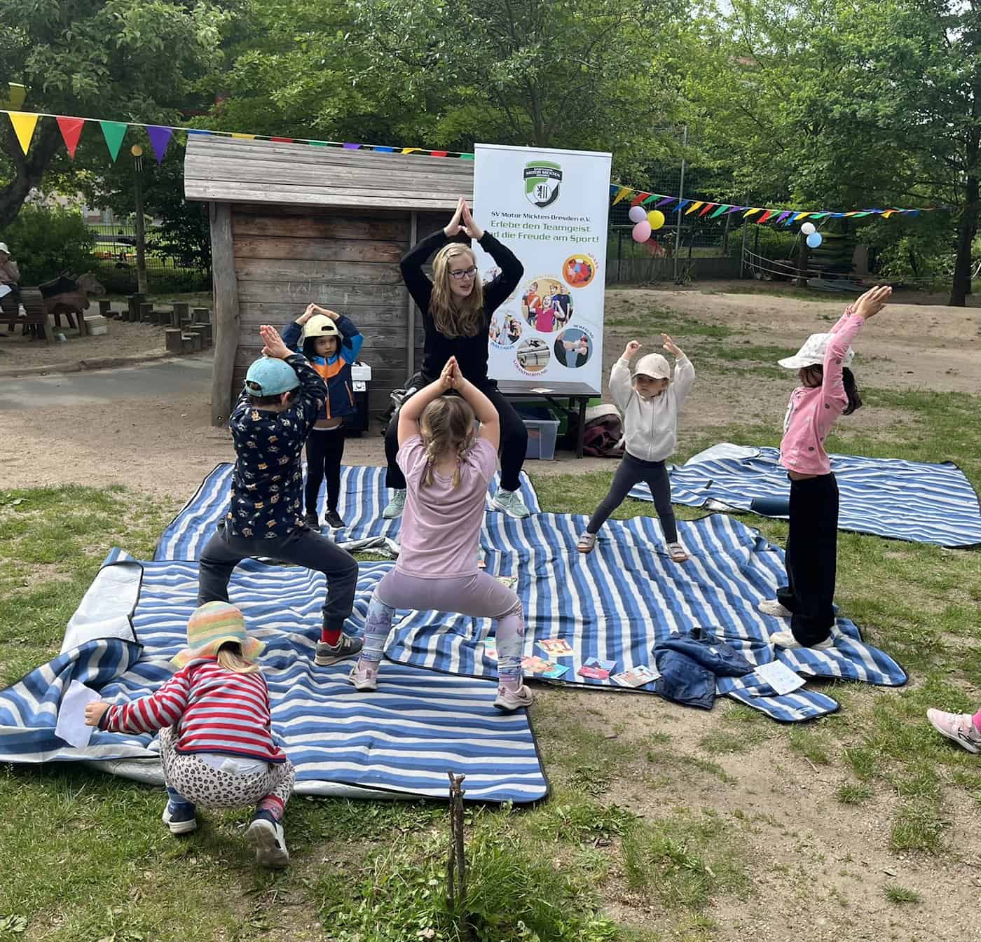 Eine Trainerin macht im Freien auf einer blau-weiß gestreiften Decke gemeinsam mit mehreren Kindern Yoga-Übungen vor einem Banner des SV Motor Mickten Dresden e.V.