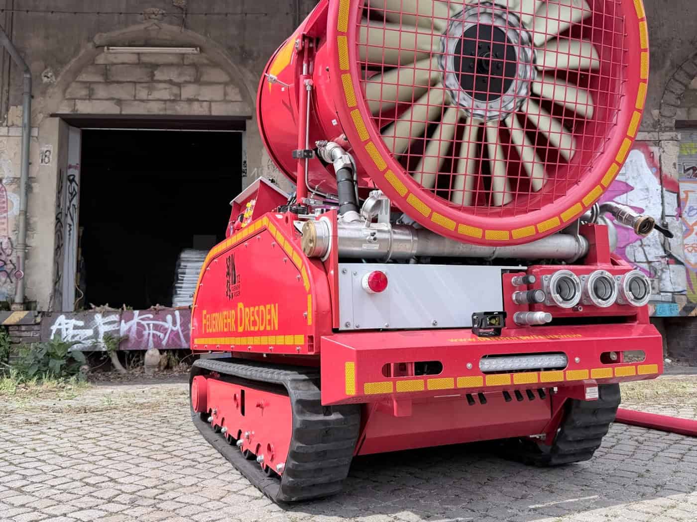 Ein spezialisiertes, ferngesteuertes Löschunterstützungsfahrzeug (Löschroboter) auf Raupenketten steht im Vordergrund-links vor einem historischen Backsteingebäude des ehemaligen Leipziger Bahnhofs in Dresden. Das robuste, unbemannte Fahrzeug hat weiße Abdeckungen über seinen Werkzeugen und steht in