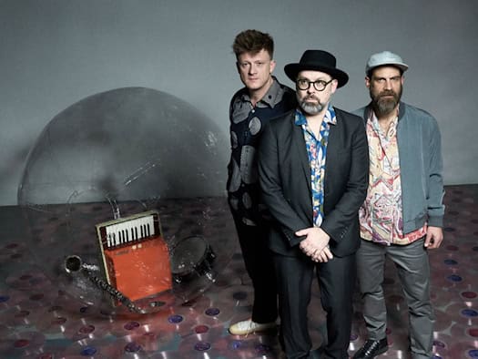 PR-Foto im Halbdunkel mit stimmungsvollem bläulichem Licht. Ein Musiker mit Brille und Bart spielt konzentriert auf einem Wurlitzer-E-Piano im Vordergrund. Links hinten steht ein weiterer Musiker mit einem Blasinstrument, während rechts im Hintergrund ein Schlagzeuger hinter seinem Set zu erken
