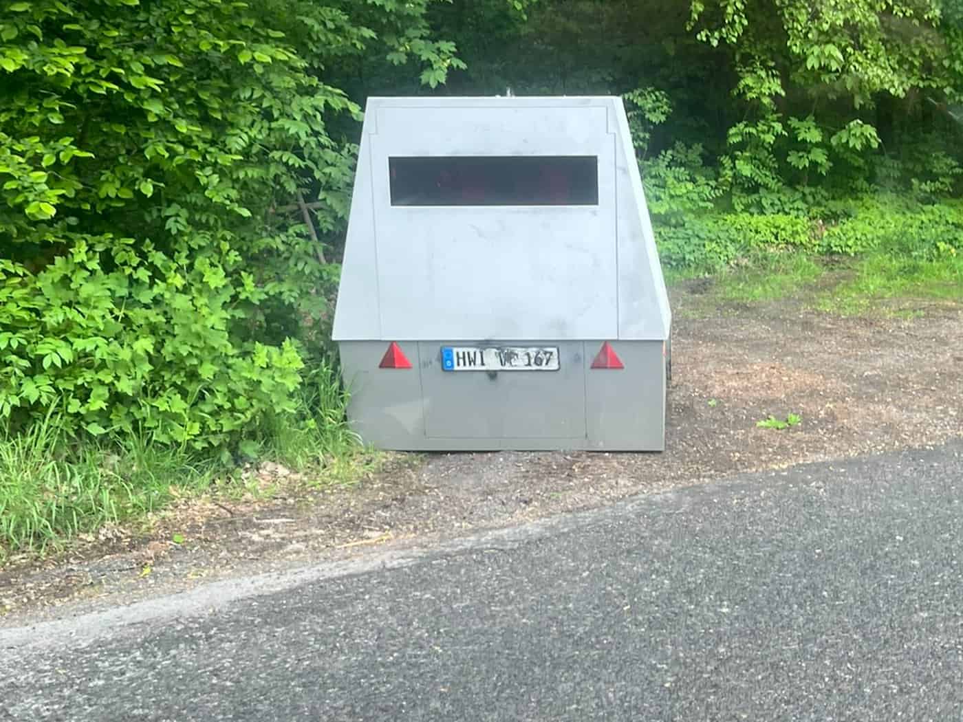 Nahaufnahme eines mobilen Geschwindigkeitsmessgeräts (Enforcement Trailer) mit dem Kennzeichen HWI-VE 167. Das Gerät ist mit Graffiti beschmiert und steht auf einem gepflasterten Gehwegabschnitt vor einem Zaun und dichten grünen Hecken.