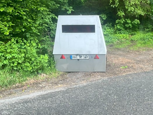 Nahaufnahme eines mobilen Geschwindigkeitsmessgeräts (Enforcement Trailer) mit dem Kennzeichen HWI-VE 167. Das Gerät ist mit Graffiti beschmiert und steht auf einem gepflasterten Gehwegabschnitt vor einem Zaun und dichten grünen Hecken.
