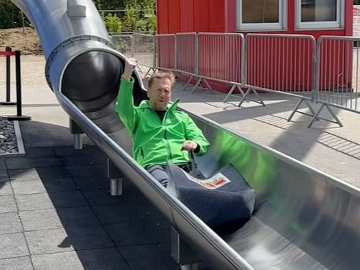 Der sächsische Ministerpräsident Michael Kretschmer rutscht lächelnd und in Freizeitkleidung durch eine geschlossene Röhrenrutsche hinunter; das Ende der Rutsche befindet sich in einer gepflegten Parkanlage.