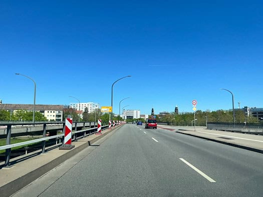 Blick über die Fahrbahn einer Brücke in Dresden bei sonnigem Wetter. Die Straße ist durch rot-weiße Warnbaken begrenzt, im Hintergrund sind Stadtgebäude und die Türme der Altstadt unter blauem Himmel zu sehen.