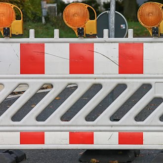 Symbolbild einer Straßensperrung: Eine weiß-rot gestreifte Absperrschranke mit drei gelben Warnleuchten steht quer auf einer asphaltierten Straße.