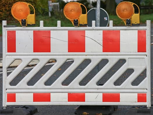 Symbolbild einer Straßensperrung: Eine weiß-rot gestreifte Absperrschranke mit drei gelben Warnleuchten steht quer auf einer asphaltierten Straße.