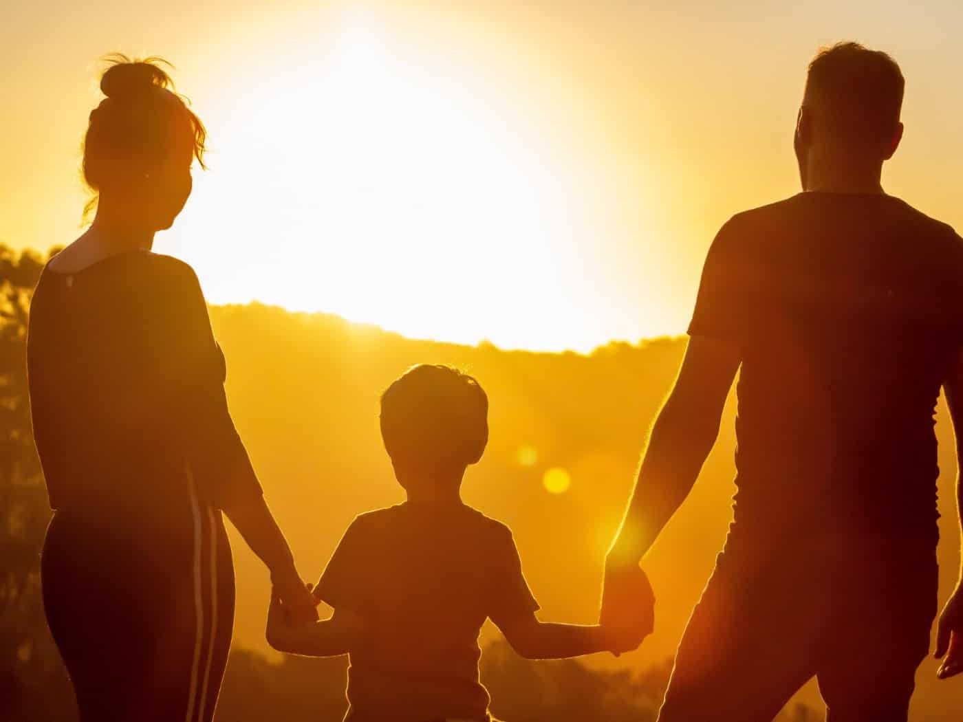 Die Silhouette einer dreiköpfigen Familie – Mutter, Vater und ein kleines Kind in der Mitte – steht Hand in Hand vor einer untergehenden Sonne. Das warme Gegenlicht erzeugt eine emotionale Atmosphäre und hebt die Umrisse der Gruppe vor einer Hügellandschaft hervor.