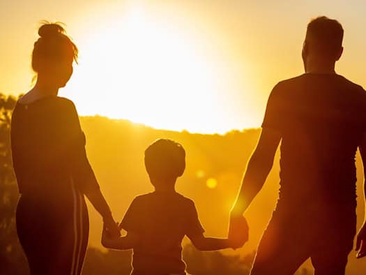 Die Silhouette einer dreiköpfigen Familie – Mutter, Vater und ein kleines Kind in der Mitte – steht Hand in Hand vor einer untergehenden Sonne. Das warme Gegenlicht erzeugt eine emotionale Atmosphäre und hebt die Umrisse der Gruppe vor einer Hügellandschaft hervor.