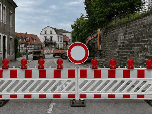 Eine rot-weiße Absperrschranke mit roten Warnleuchten und einem runden Durchfahrtsverbotsschild blockiert die Straße in Dresden-Wachwitz. Im Hintergrund sind ein gelber Baubagger und eine historische Bruchsteinmauer zu sehen.