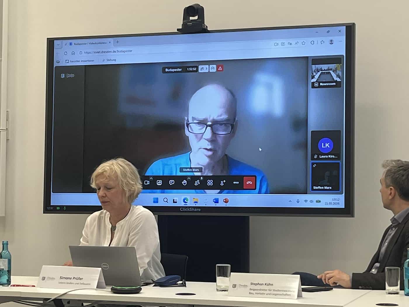 Tiefbauamtsleiterin Simone Prüfer und Baubürgermeister Stephan Kühn bei einer Pressekonferenz an einem Tisch. Auf einem großen Bildschirm im Hintergrund ist Professor Steffen Marx per Video zugeschaltet.