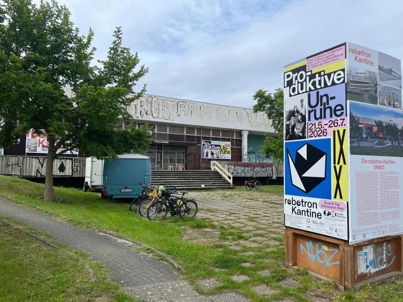Die denkmalgeschützte robotron-Kantine in Dresden, davor parkende Fahrräder und eine Infostele zur Ausstellung „Produktive Unruhe“.