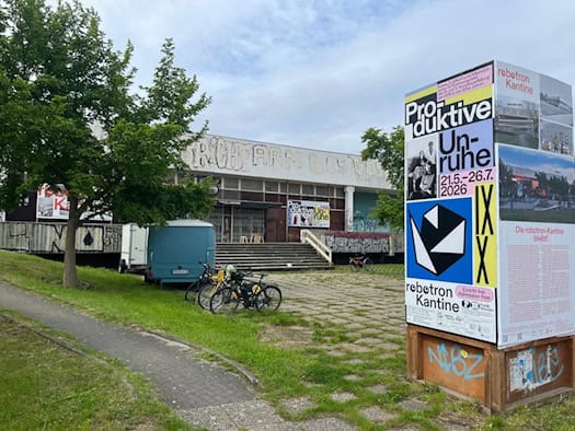 Die denkmalgeschützte robotron-Kantine in Dresden, davor parkende Fahrräder und eine Infostele zur Ausstellung „Produktive Unruhe“.