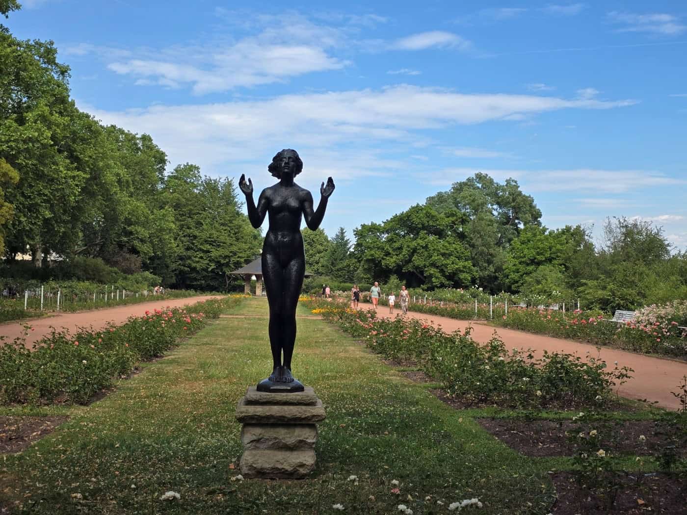 Eine von Bäumen gesäumte, symmetrische Gartenanlage mit Rosenbeeten unter einem leicht bewölkten, blauen Himmel. Im Vordergrund steht die dunkle Bronzeplastik einer nackten Frau mit erhobenen Händen auf einem mehrstufigen Steinsockel. Im Hintergrund flanieren einige Spaziergänger auf dem zentralen S