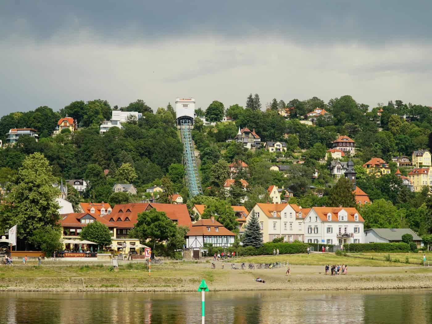 Weitwinkelaufnahme der Elbe im Vordergrund, dahinter die Wohnhäuser von Loschwitz und die steil den Hang hinauführende Trasse der Dresdner Schwebebahn unter einem bewölkten Himmel.