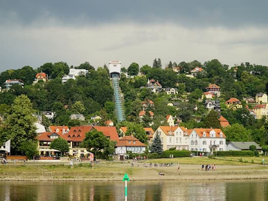 Weitwinkelaufnahme der Elbe im Vordergrund, dahinter die Wohnhäuser von Loschwitz und die steil den Hang hinauführende Trasse der Dresdner Schwebebahn unter einem bewölkten Himmel.
