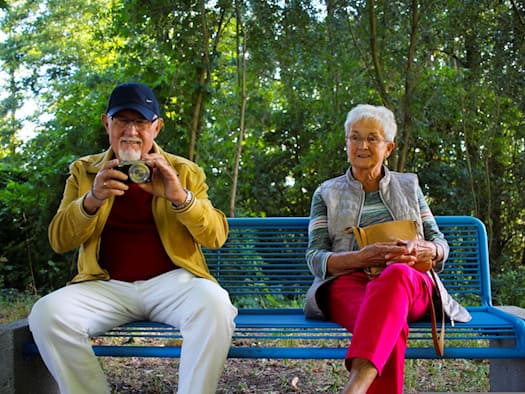 Ein älteres Paar sitzt gemeinsam entspannt auf einer blauen Parkbank in einem grünen Park; der Mann hält eine Kamera in den Händen.