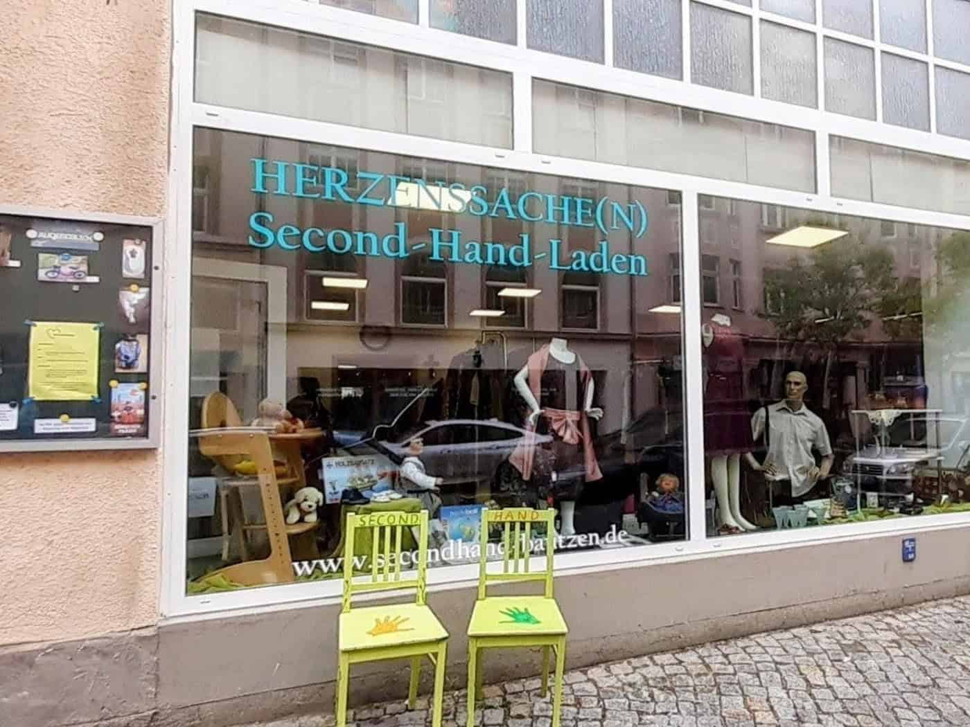 Schaufenster des Second-Hand-Ladens Herzenssache(n) in Bautzen mit Schaufenster, Kleidung, Spielzeug und bunten Stühlen vor dem Eingang.